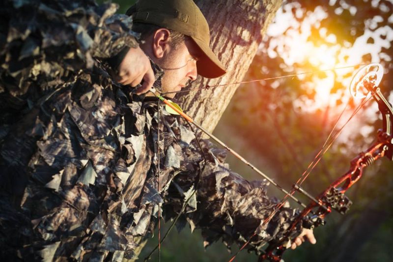 Camouflaged bowhunter aiming a quiet compound bow at sunset