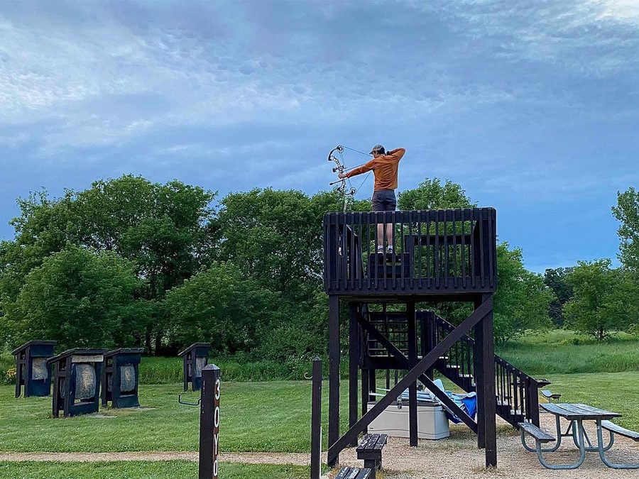 Archer at full draw shooting a compound bow from an elevated platform at a 3D archery range