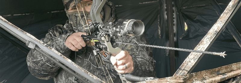 Bowhunter drawing compound bow inside ground blind through shooting window